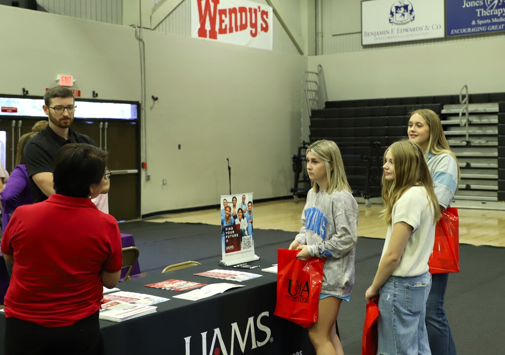 Three female students standing near a college fair booth labeled UAMS, listening attentively to college representatives at the display table.