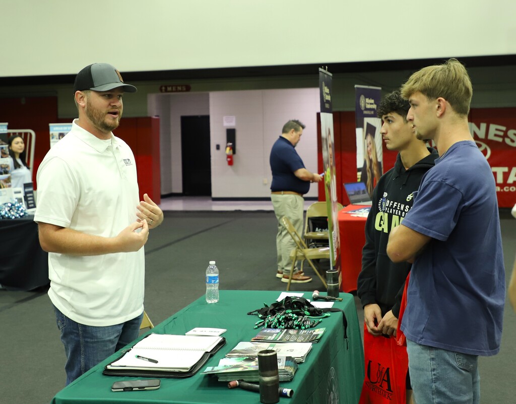 A college representative in a white polo shirt and grey cap talking to two high school students at a green table set up with informational brochures at a college fair.