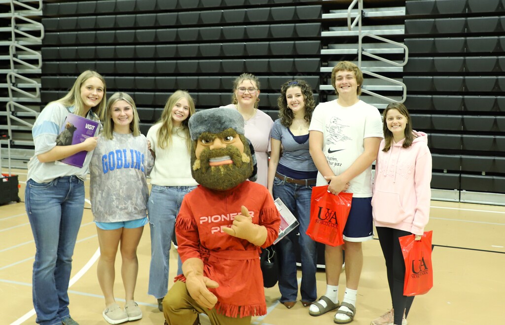 A group of seven Harrison High School students posing indoors on a gymnasium floor alongside the UofA Northark Pioneer Pete mascot in a red and fur-trimmed outfit. Some students are holding red gift bags and information folders.