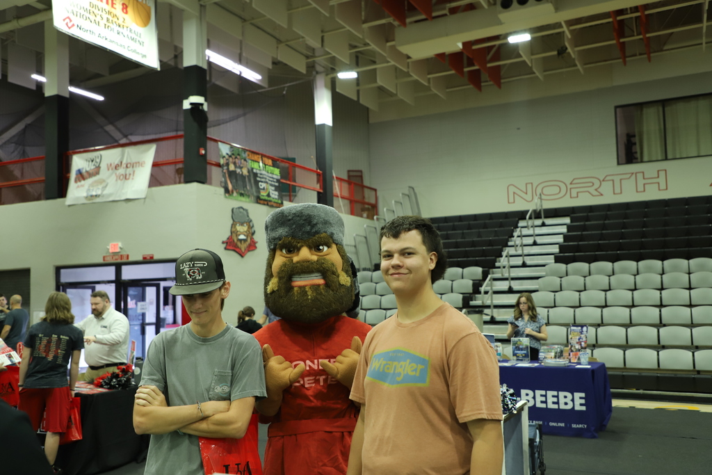 Two high school students posing for a photo on the gymnasium floor on either side of the UofA Northark Pioneer Pete mascot, with college fair tables visible in the background.