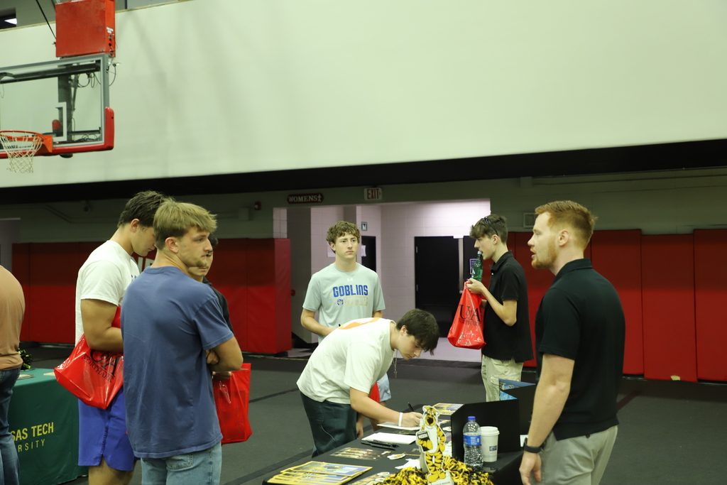 Several high school students gathered around college information tables inside a gymnasium, with one student filling out a sign-in sheet at an Arkansas Tech table.