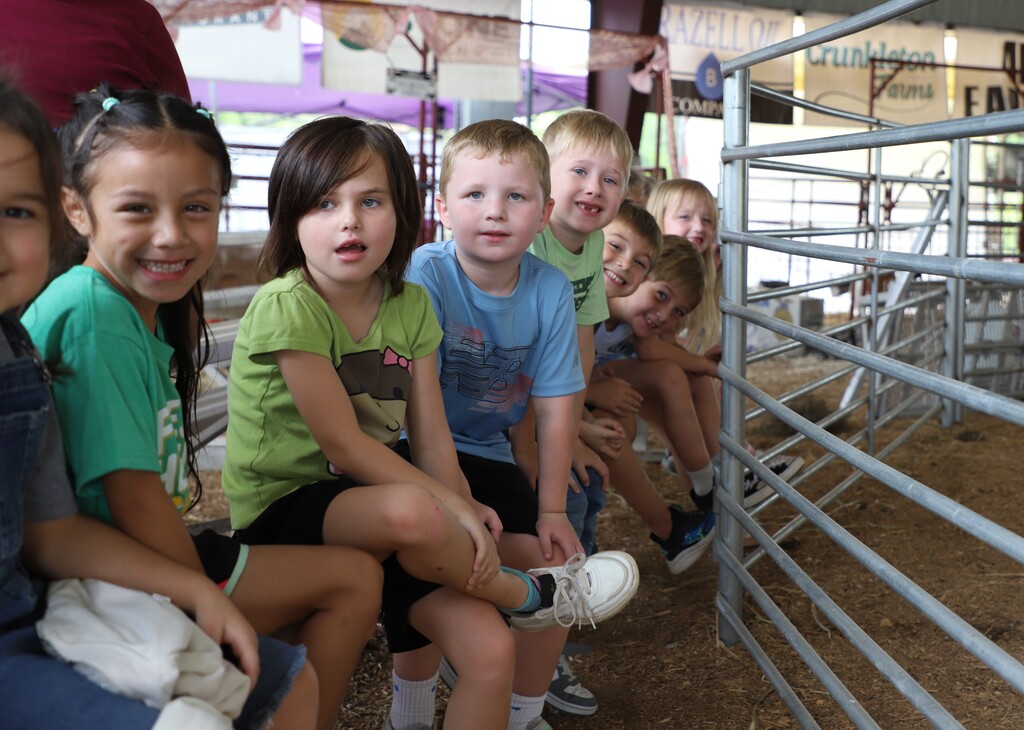 A group of elementary school children sitting together on a bench near a metal livestock pen at the fairgrounds, smiling and looking toward the camera.