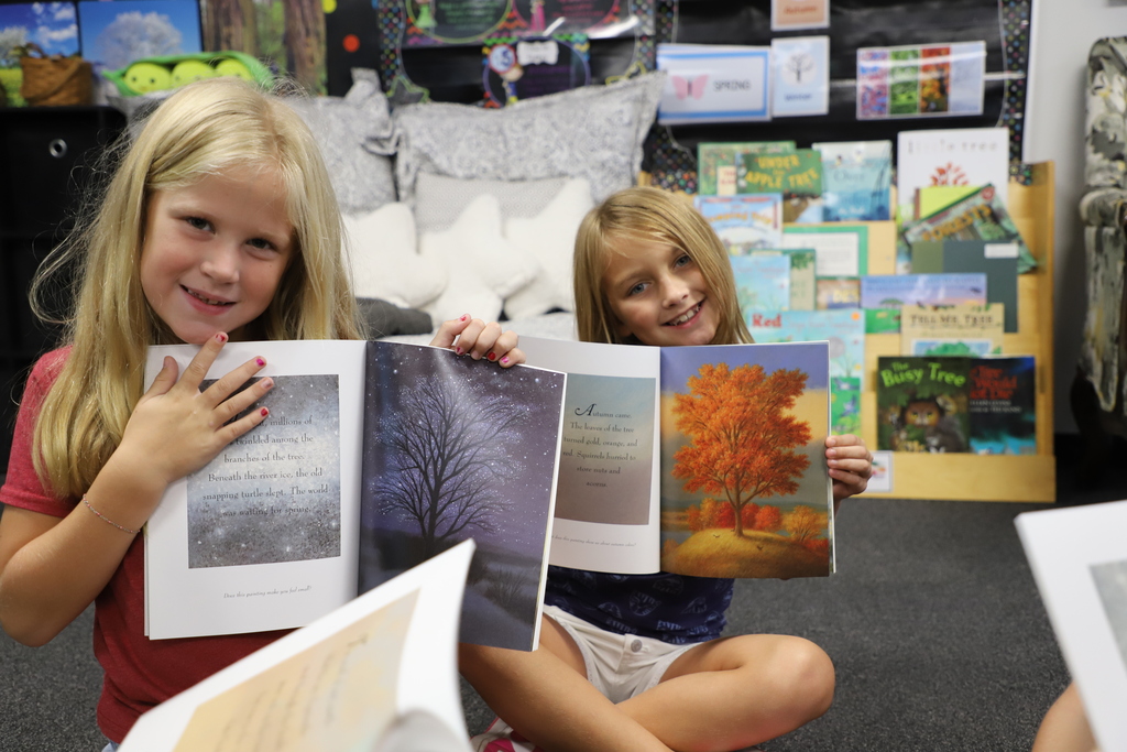 "Two young female elementary students sitting indoors and smiling while holding open picture books about fall trees and autumn landscapes."