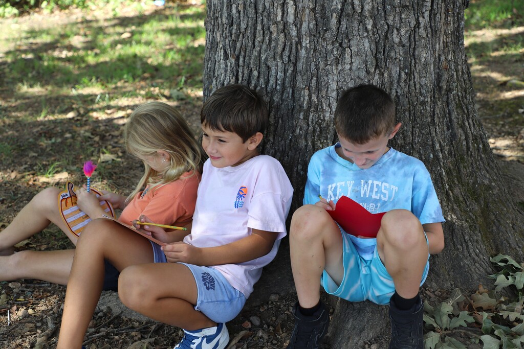 "Three children sitting outdoors at the base of a large tree trunk, reading and writing on colorful fall-themed paper cutouts."