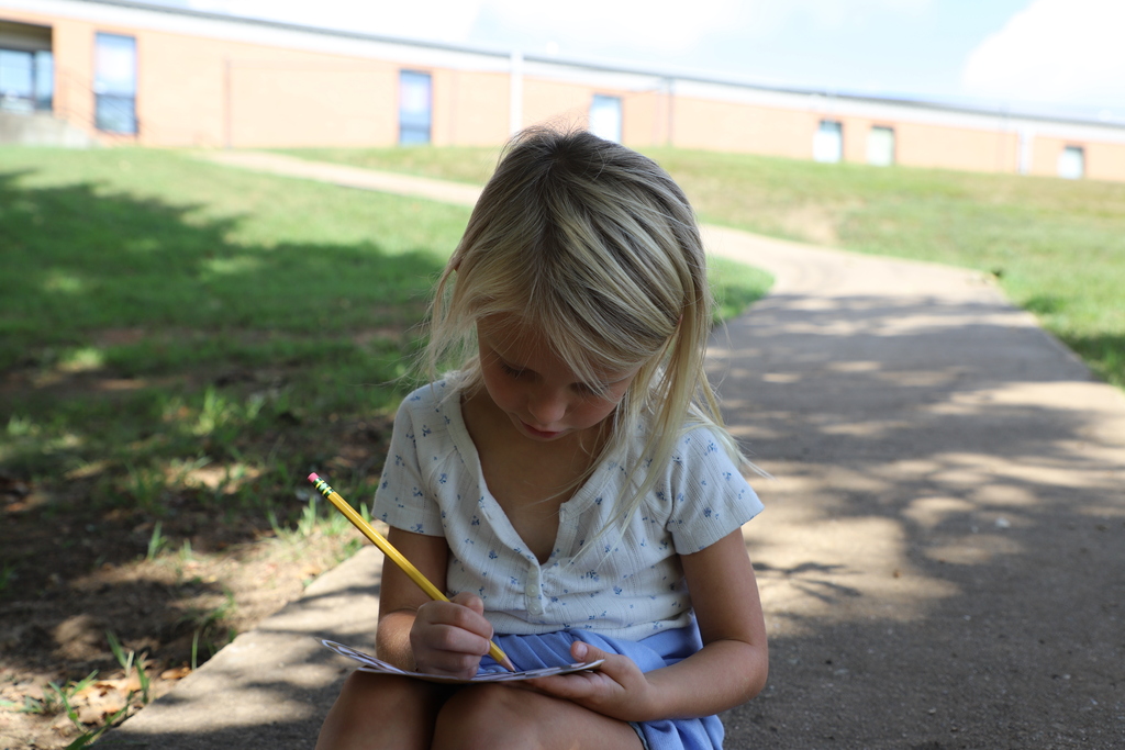 "A young girl sitting outside on a sidewalk path, focusing intently on writing with a yellow pencil on paper."