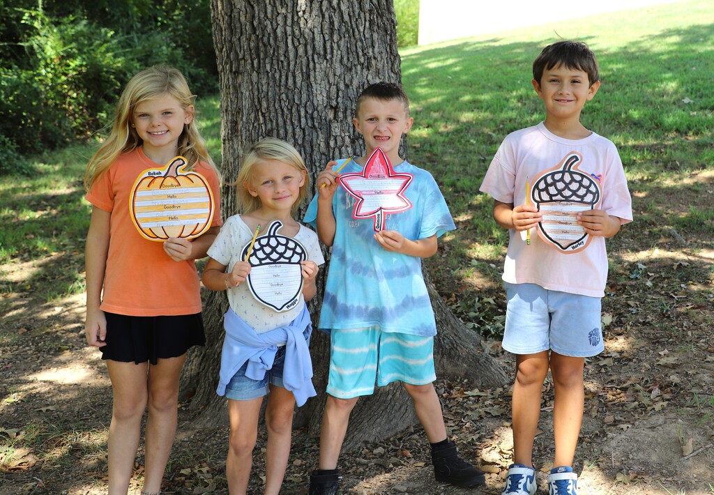 "Four smiling elementary students standing outdoors in front of a tree, holding up fall-themed paper cutouts shaped like pumpkins, acorns, and leaves."