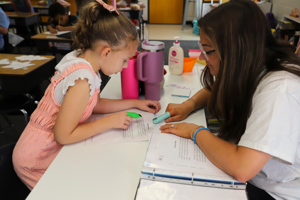 "A teacher and a young female student sitting at a desk together, using green pointing 'monster fingers' to read a printed passage."