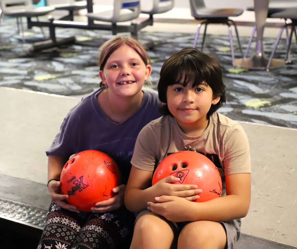 Two young elementary students sitting together on the floor of a bowling alley, smiling while hugging orange bowling balls.