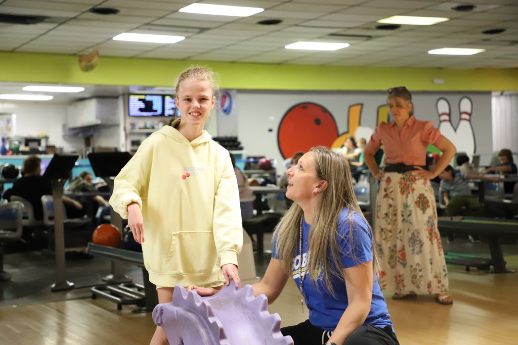 : A young female student in a yellow hoodie standing at a bowling lane next to a purple ramp, with a smiling staff member kneeling beside her to assist.