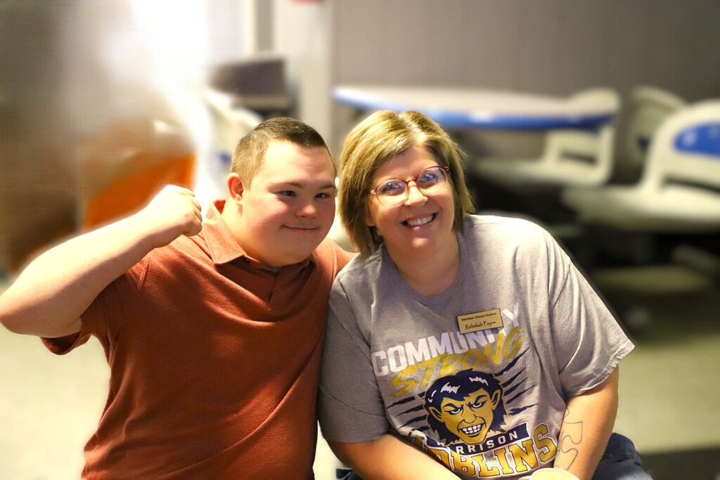 A smiling young male student in a terracotta polo shirt flexing his arm next to a smiling female staff member wearing a gray Harrison Goblins shirt and glasses.