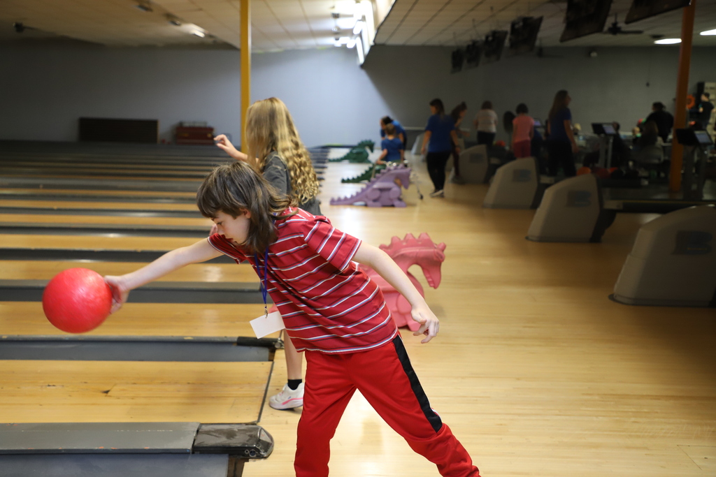 A student in a red striped shirt leaning forward on a bowling lane, about to roll a red bowling ball down the alley.