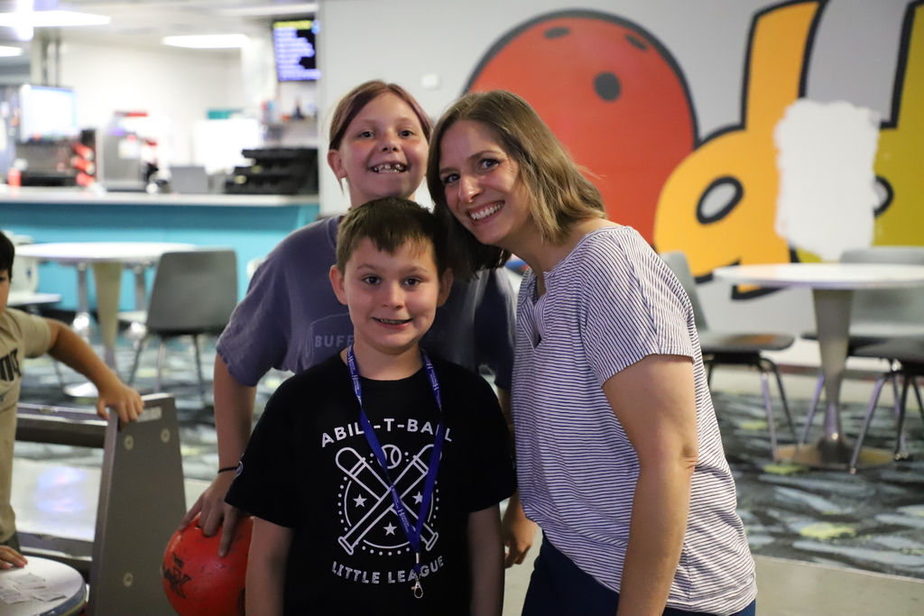 A young boy wearing a black "Abili-T-Ball" t-shirt and lanyard standing with a girl and a female staff member smiling for the camera at a bowling alley.