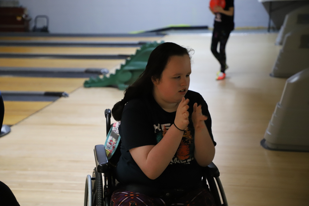 A female student in a black t-shirt sitting in a wheelchair and clapping her hands with a focused smile at the bowling alley.