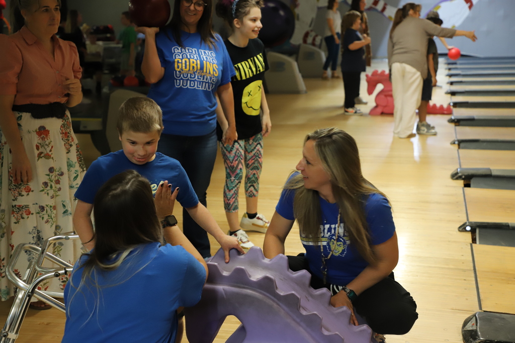 A young male student giving a high-five to a staff member kneeling next to a purple bowling ramp, surrounded by cheering classmates and aides.