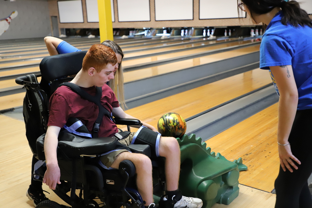 A male student in a wheelchair positioned at the top of a green bowling ramp, preparing to launch a marbleized bowling ball with the help of a staff member.