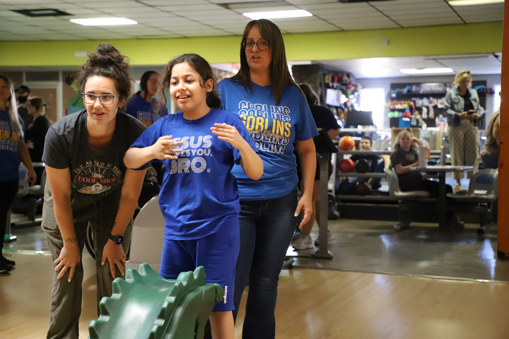 A girl wearing a blue t-shirt guiding a bowling ball down a green dragon-shaped ramp, supported by two female staff members kneeling beside her.