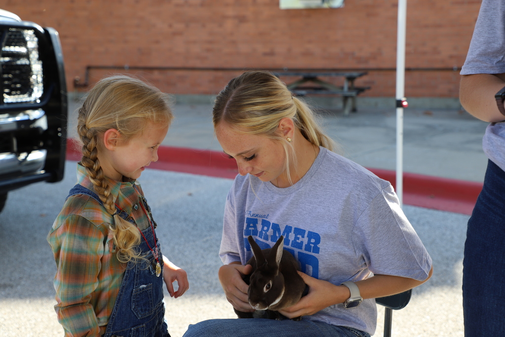 A high school student wearing a grey "Farmer Fred Day" shirt sits with a brown rabbit in her lap while a young girl in overalls and a plaid shirt looks on with a gentle smile.