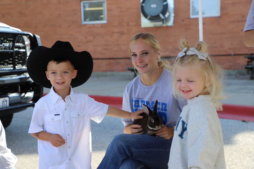 A young boy in a black cowboy hat and white button-down shirt touches a brown rabbit held by a high school student, while a young girl in a grey sweater smiles beside them outdoors.