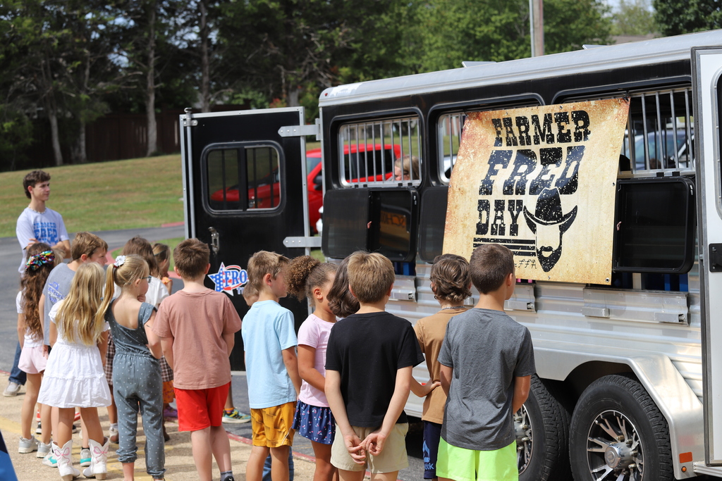 A line of elementary students waiting outside near a livestock trailer adorned with a "Farmer Fred Days" sign.