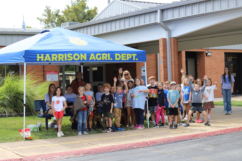 A large group of elementary school students and high school volunteers smiling and waving for a group photo under a blue tent that reads "Harrison Agri. Dept." in front of the school building.