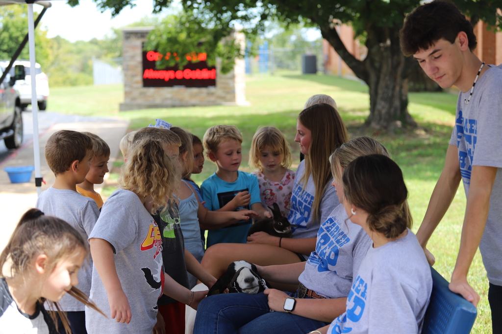 High school volunteers sitting outdoors holding a brown rabbit and a goat while a group of curious elementary students gather around to pet them.