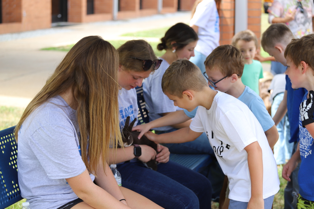 High school students sitting on an outdoor bench holding a small rabbit while elementary school children lean in to gently pet it.