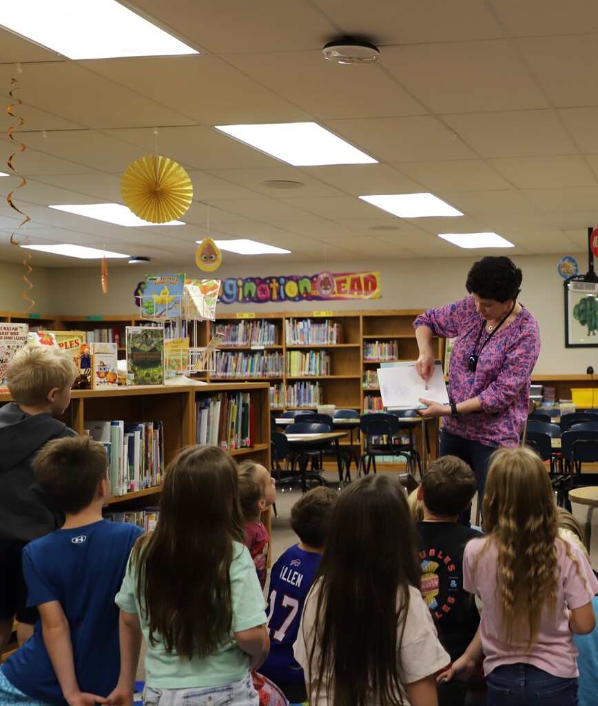 A wide shot of first-grade students standing together on the rug in the school library, looking up closely as Mrs. Ward demonstrates a paper craft activity holding up a sheet of paper.