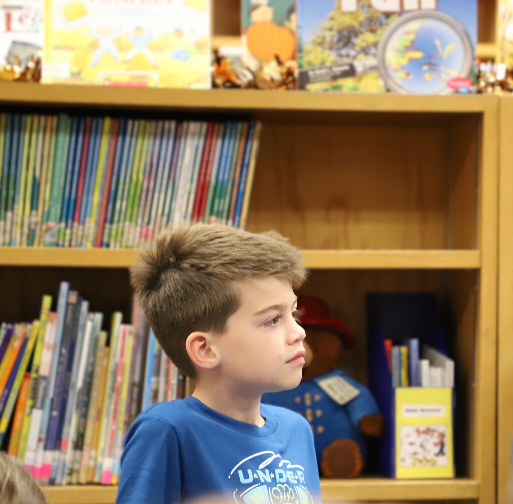 A young schoolboy in a blue shirt standing up attentively in the library during storytime, looking off-camera toward the librarian while listening or preparing to ask a question.