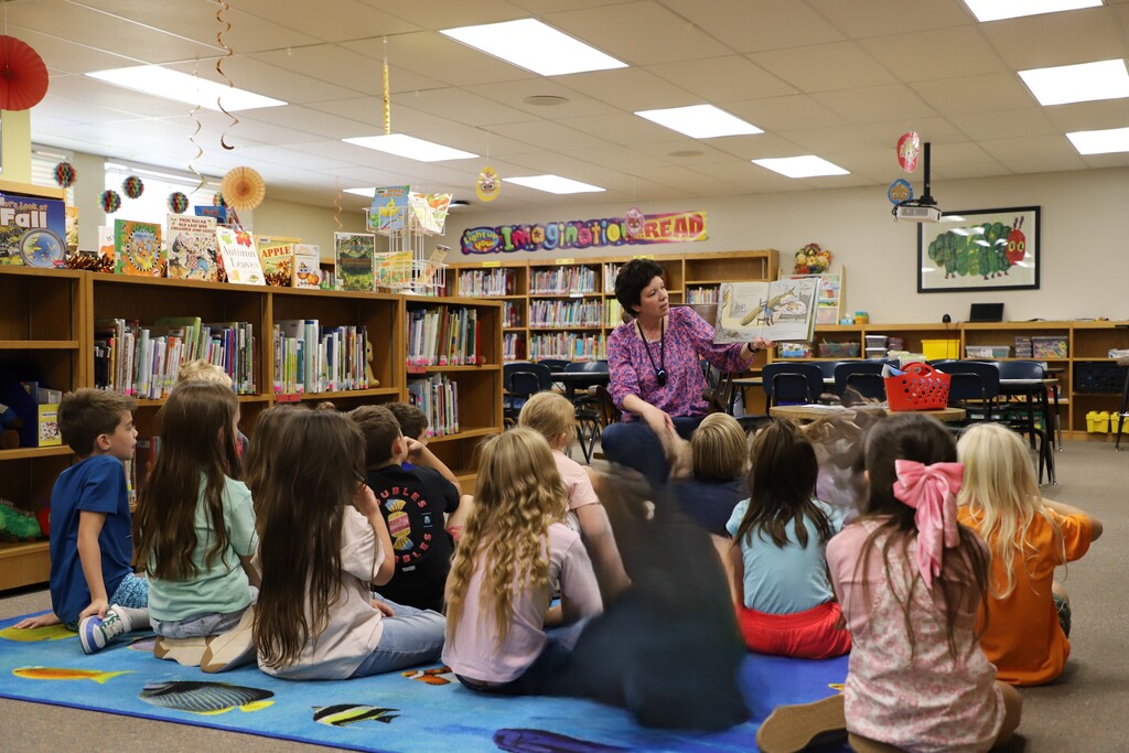 A group of first-grade elementary students sitting cross-legged on a colorful fish-themed rug in the school library, attentively listening to librarian Mrs. Ward as she sits in front of them holding up an open picture book.