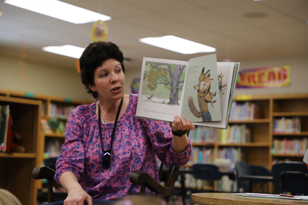 Close-up of librarian Mrs. Ward sitting in the library, smiling as she holds open the picture book Lawrence in the Fall to display the colorful illustrations to her class.