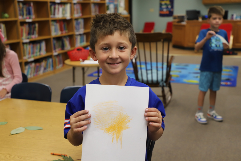 A young boy smiling proudly at the camera while holding up his completed yellow leaf rubbing artwork on a white piece of paper.