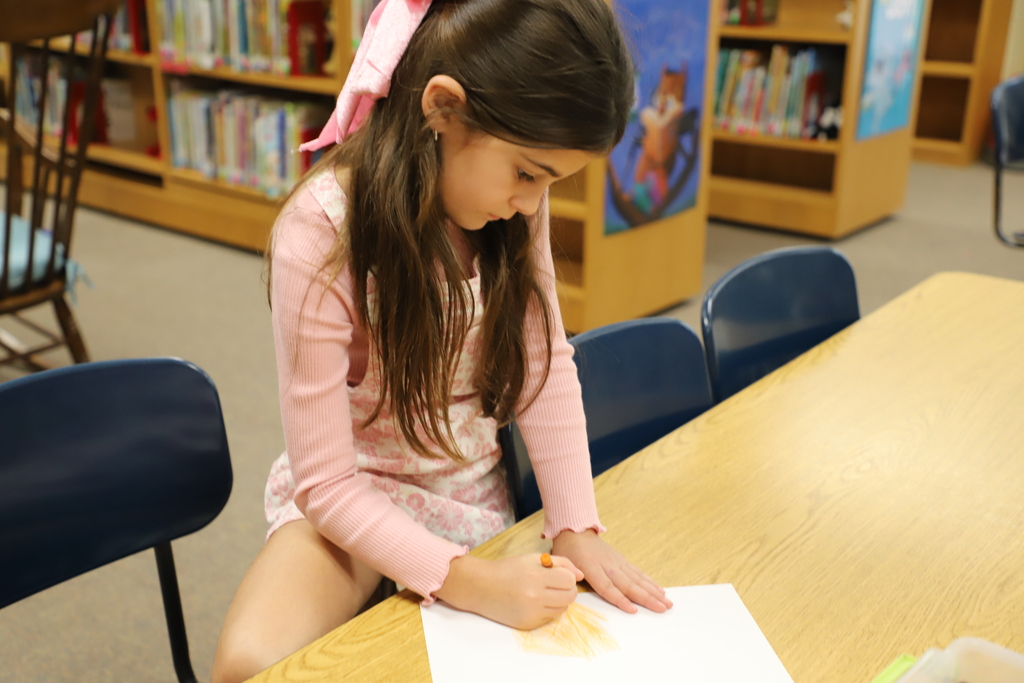 A young girl with a pink bow in her hair sitting at a wooden library table, using a crayon to carefully create a leaf rubbing on a white sheet of paper.