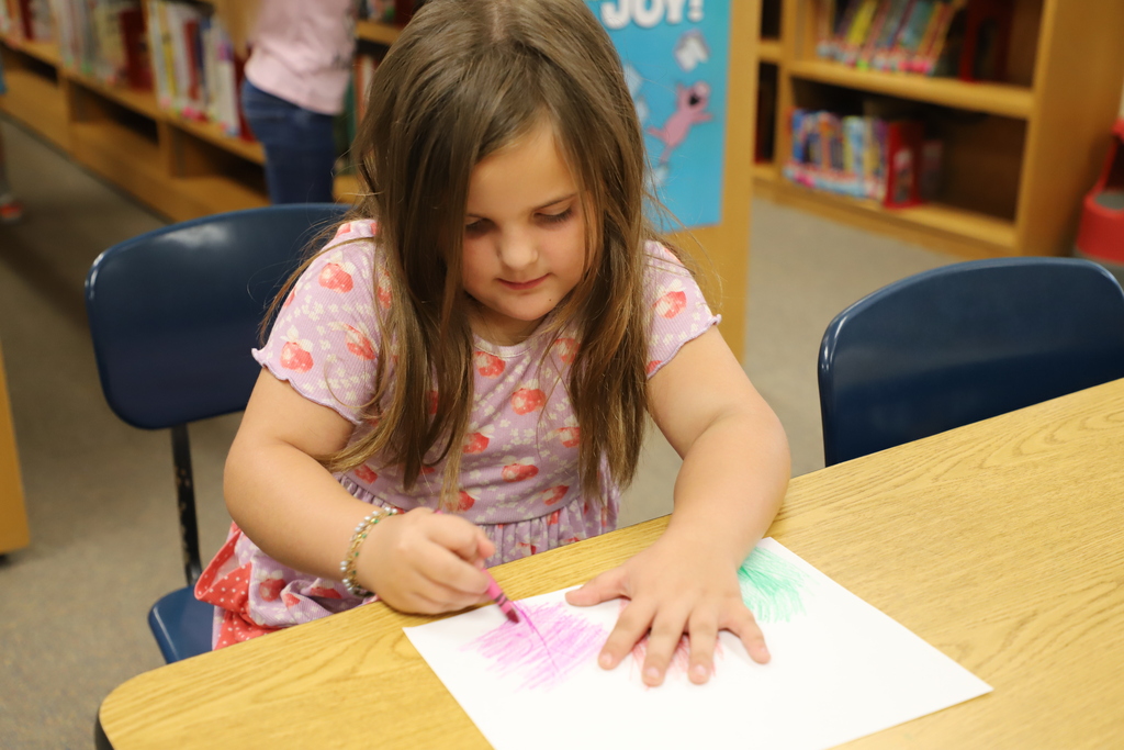 A young girl in a pink patterned dress concentrating as she rubs a crayon over a leaf placed under white paper at a classroom table.