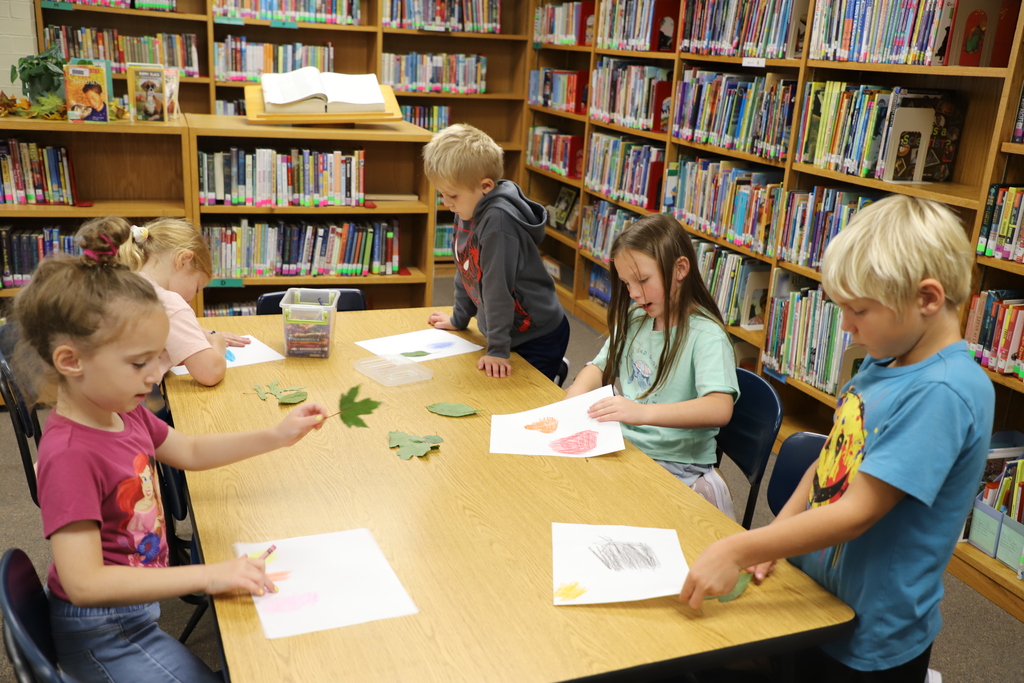 Several elementary students gathered around a long wooden table in the library, working together with crayons and real leaves to make leaf rubbings.