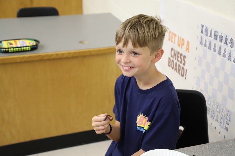 A smiling young boy with short blonde hair sits at a classroom table, holding a small brown earthworm between his fingers.
