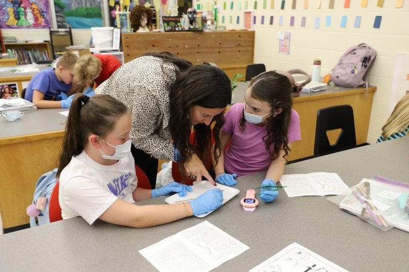 A teacher bends over a classroom desk to guide two young students wearing face masks and blue gloves as they examine a dissection tray together.