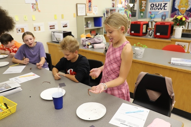 A young girl in a pink plaid dress stands in a classroom, playfully sticking out her tongue while holding a live earthworm in her palm over a table where two other classmates observe.