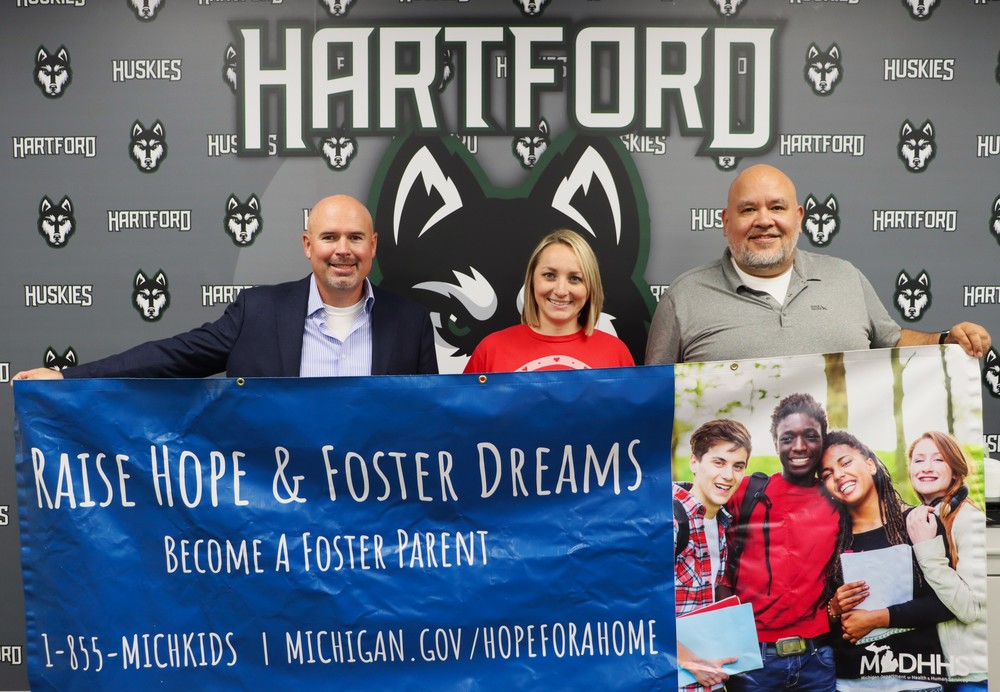 superintendent nick blackmer, athletic director peter aranda, and family bound rep casey smith pose holding a banner that reads raise hope and foster dream. become a foster parent, infront of the hartford husky logo backdrop