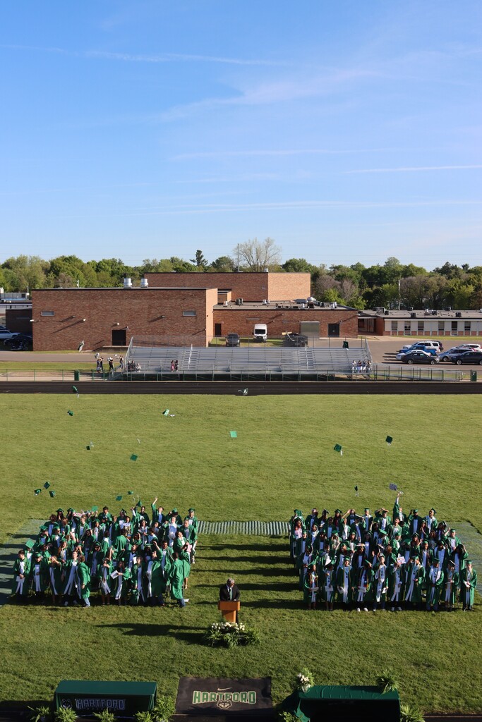Aerial view of Hartford High School’s graduation ceremony on the football field. Graduates wearing green caps and gowns stand in groups on the grass while tossing caps into the air in celebration. A speaker stands at a podium in the center of the field, with Hartford-branded tables and decorations in the foreground. Bleachers, school buildings, parked cars, and a clear blue sky appear in the background.