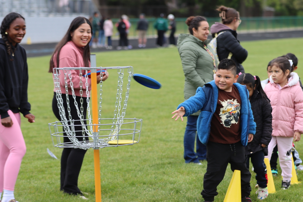A young boy throws a blue disc toward a disc golf basket during Field Day while high school students and younger kids smile and watch on the grass field. The scene captures a fun, energetic atmosphere with teamwork, games, and school spirit.
