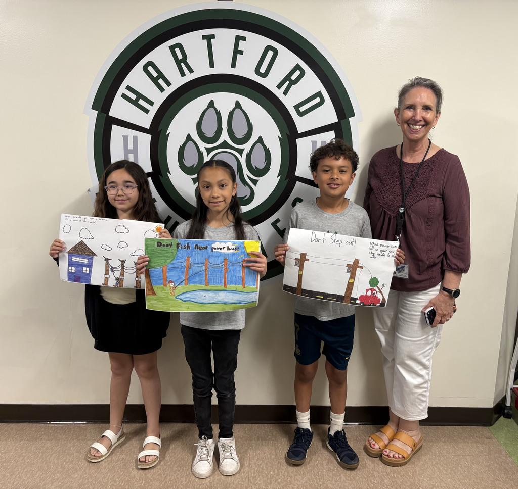 Three children and one adult are posing together. The children are holding colorful safety-themed drawings: One poster warns about staying away from power lines near homes. One says “Don’t Fish Near power lines!” Another says “Don’t Step out!” with a drawing of a car and fallen power lines. The adult appears to be a teacher or school staff member wearing a lanyard.