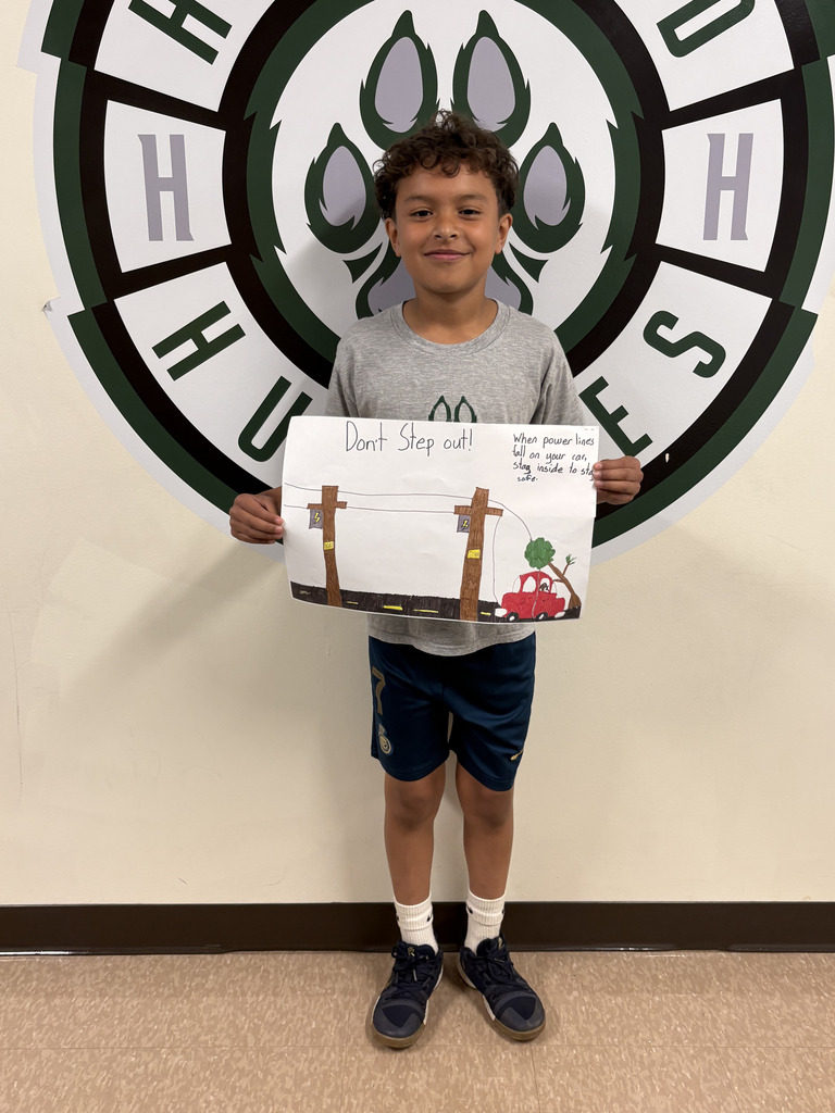 A young boy is holding a poster showing utility poles, power lines, and a red car near a fallen line. The poster says: “Don’t Step out!” “When power lines fall on your car, stay inside to stay safe.” He is also standing in front of the Hartford Huskies logo.