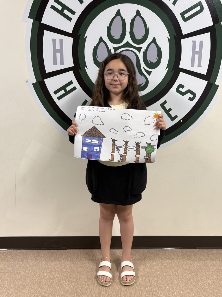 A young girl with glasses is holding a poster featuring a blue house, utility poles, and power lines. The drawing includes a warning message about staying away from power lines. She is standing in front of the Hartford Huskies logo.