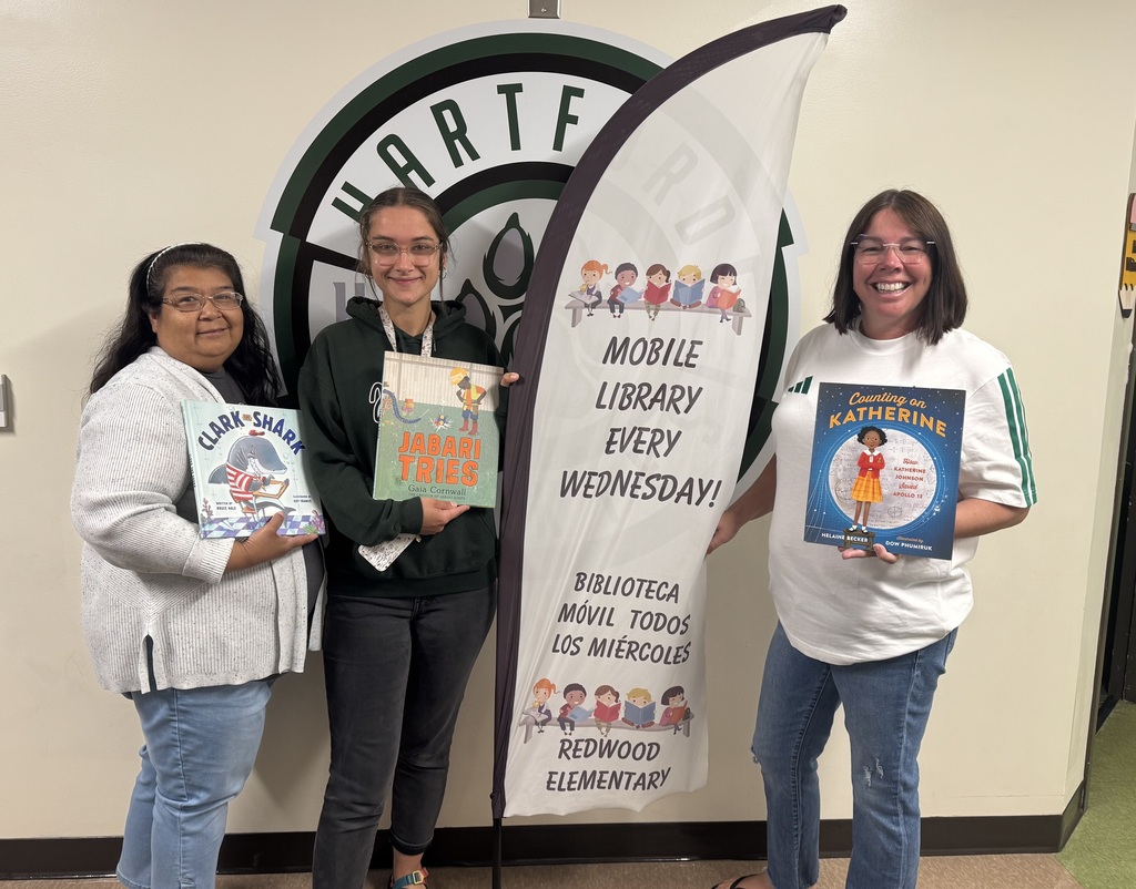 Three smiling adults stand in front of a large Redwood Elementary logo and a vertical “Mobile Library Every Wednesday” banner. Each person is holding a children’s book: Clark the Shark, Jabari Tries, and Counting on Katherine. The banner includes English and Spanish text promoting the school’s mobile library program.