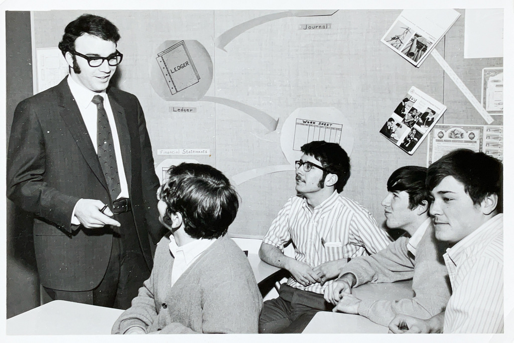 A teacher leads a classroom discussion while several high school students sit at desks listening during a business or accounting lesson.