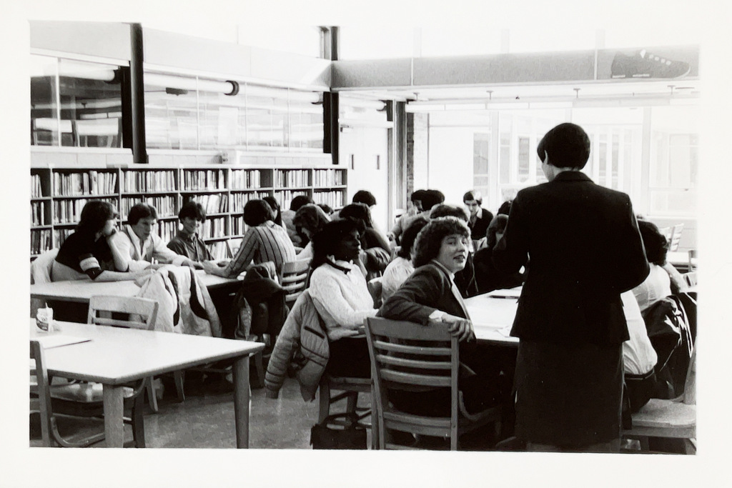 High school students study and collaborate at tables in the school library while a staff member stands nearby.