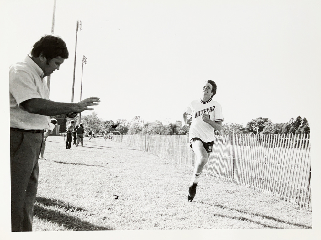 A Hartford student crosses the finish line during a cross-country meet as a race official records the time.
