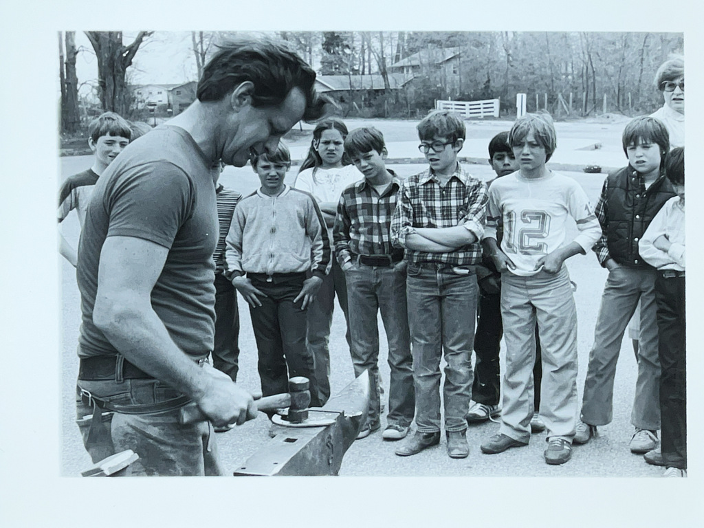 An instructor demonstrates blacksmithing techniques on an anvil while a group of students watches attentively during an outdoor hands-on lesson.