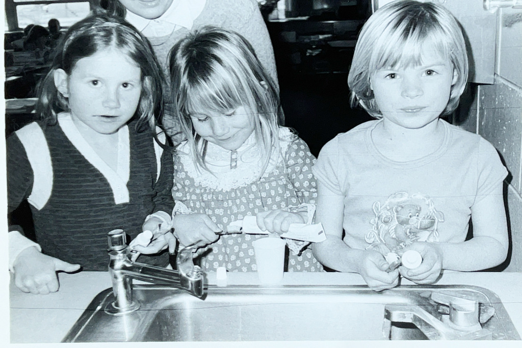 Three young elementary students gather at a classroom sink while using droppers and small containers during a science activity.