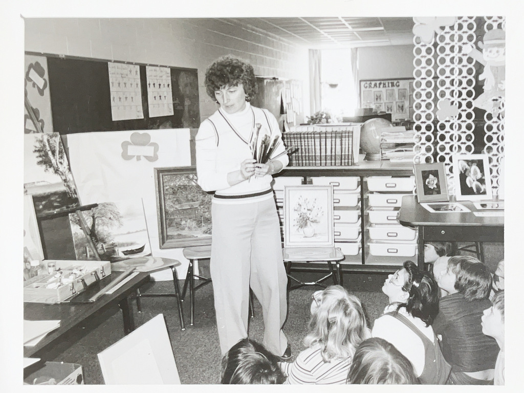 An art instructor presents paintings and brushes to a seated group of elementary students during an art demonstration in the classroom.